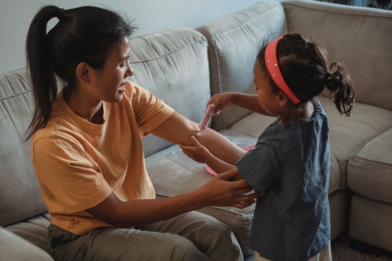 Side view of ethnic daughter making injection with fake syringe to Asian mother while playing game in living room during weekend