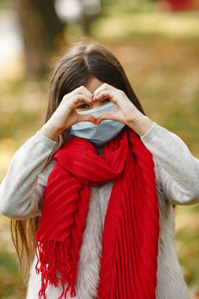 Girl in a mask and red scarf making a heart gesture during fall.