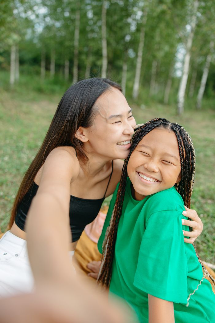 Smiling mother and daughter with braided hair taking a selfie outdoors during a summer picnic.
